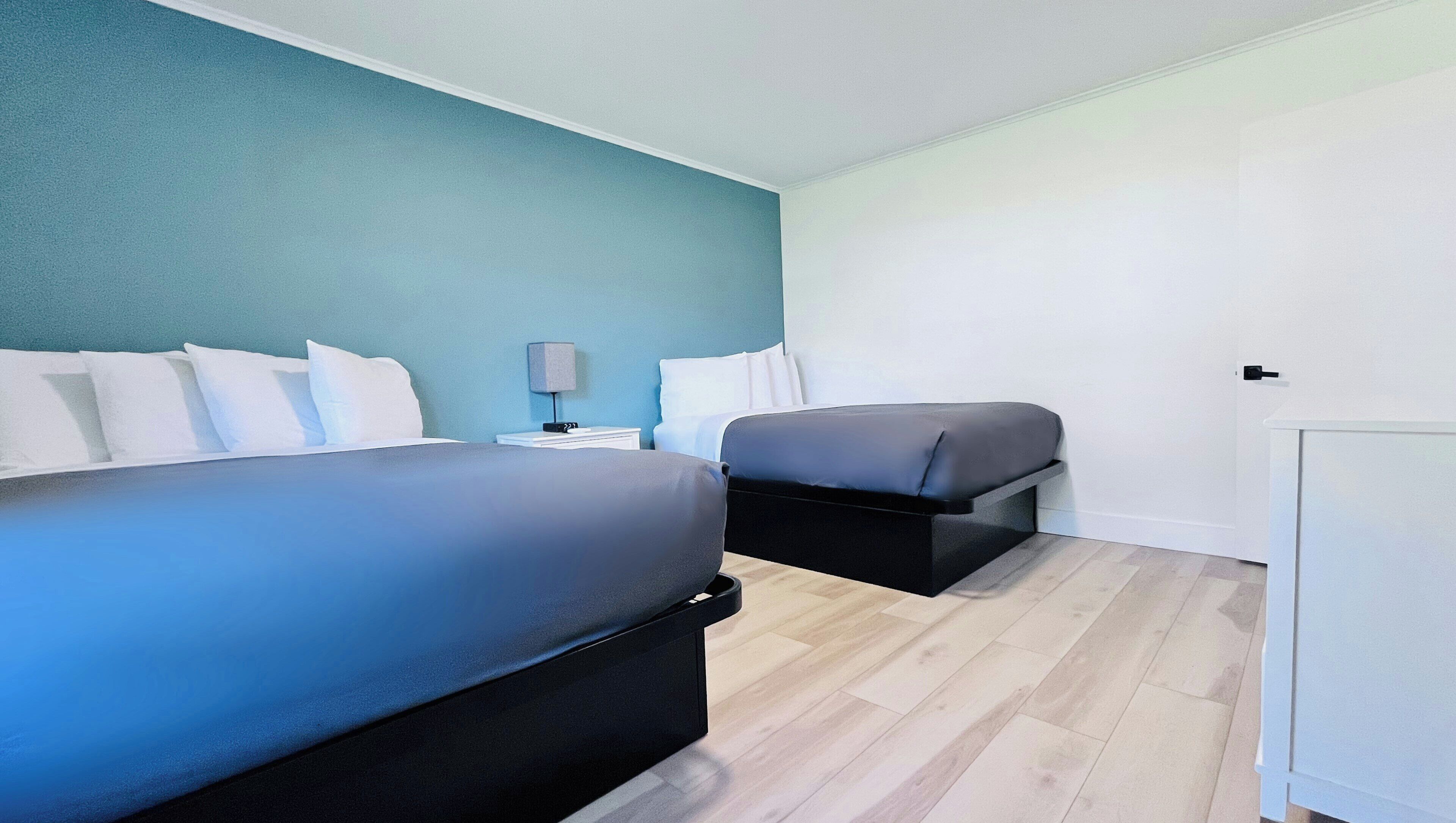 A hotel room with two beds, a bedside table with a lamp, and a white wardrobe.