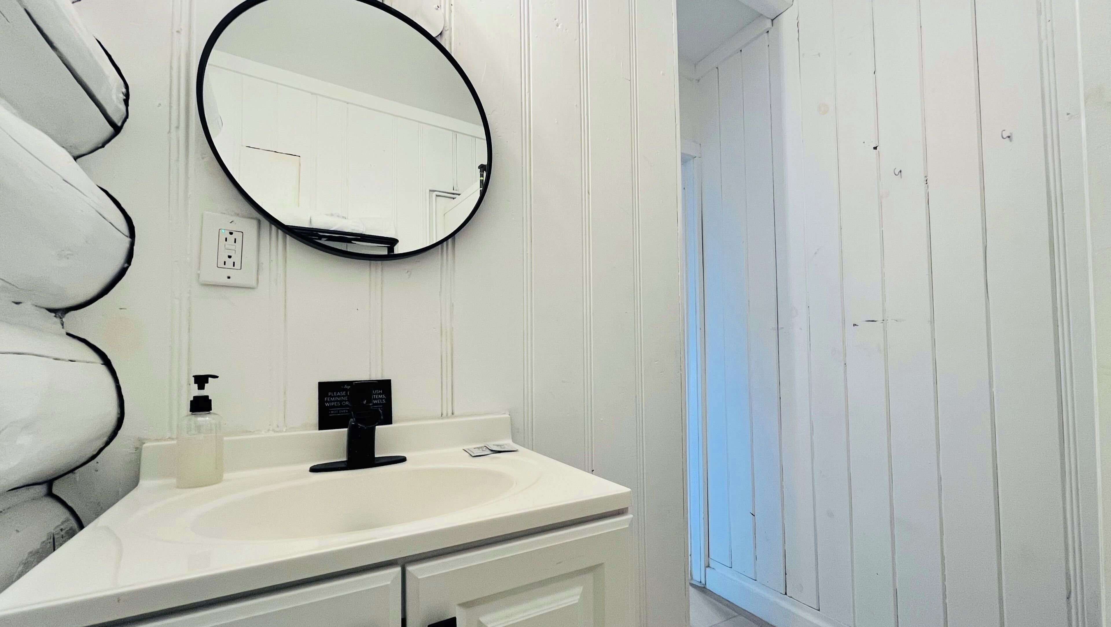 Un baño con tocador blanco, espejo redondo, grifo negro y ventana con paredes de madera blanca.
