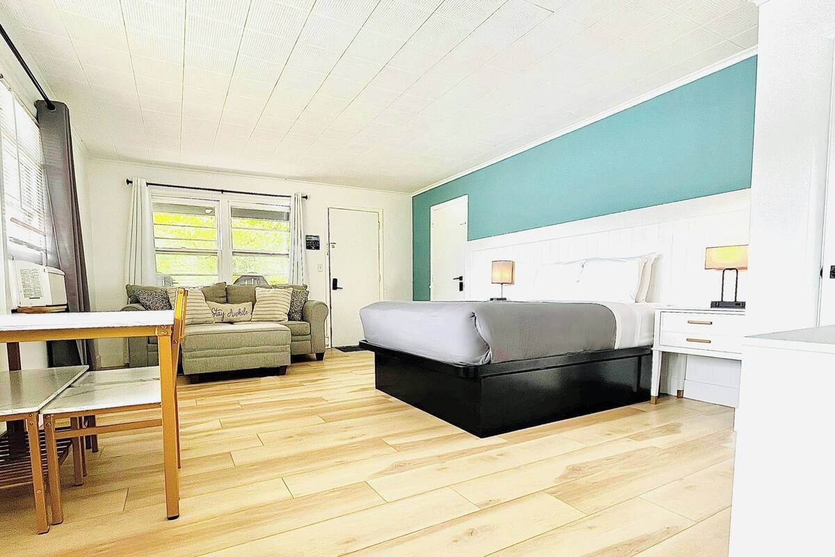 A compact room with a bed, a small table, a chair, a sofa, and a window with curtains.
