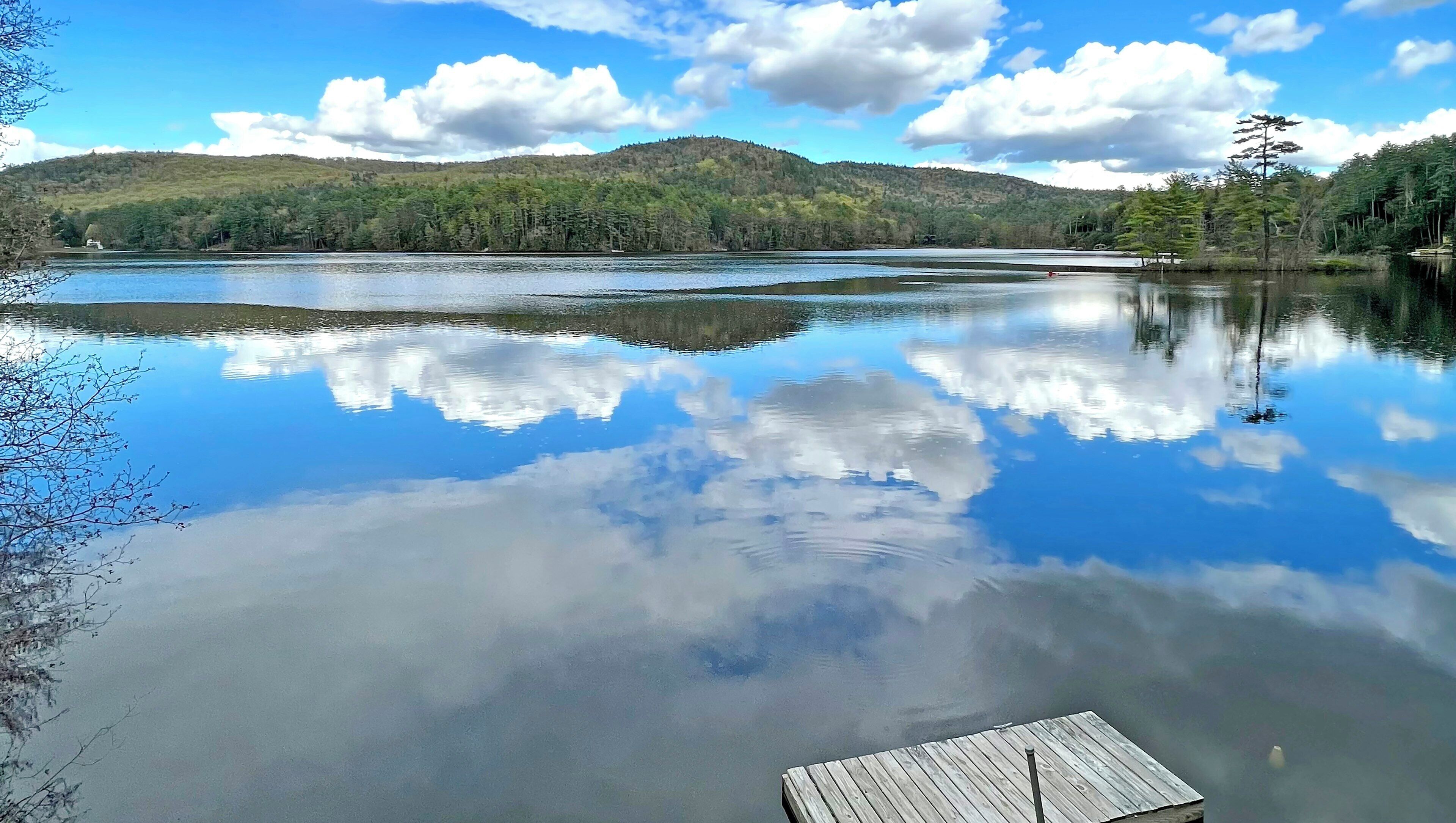 Un lago sereno con un muelle de madera, rodeado de colinas boscosas y cielos despejados.