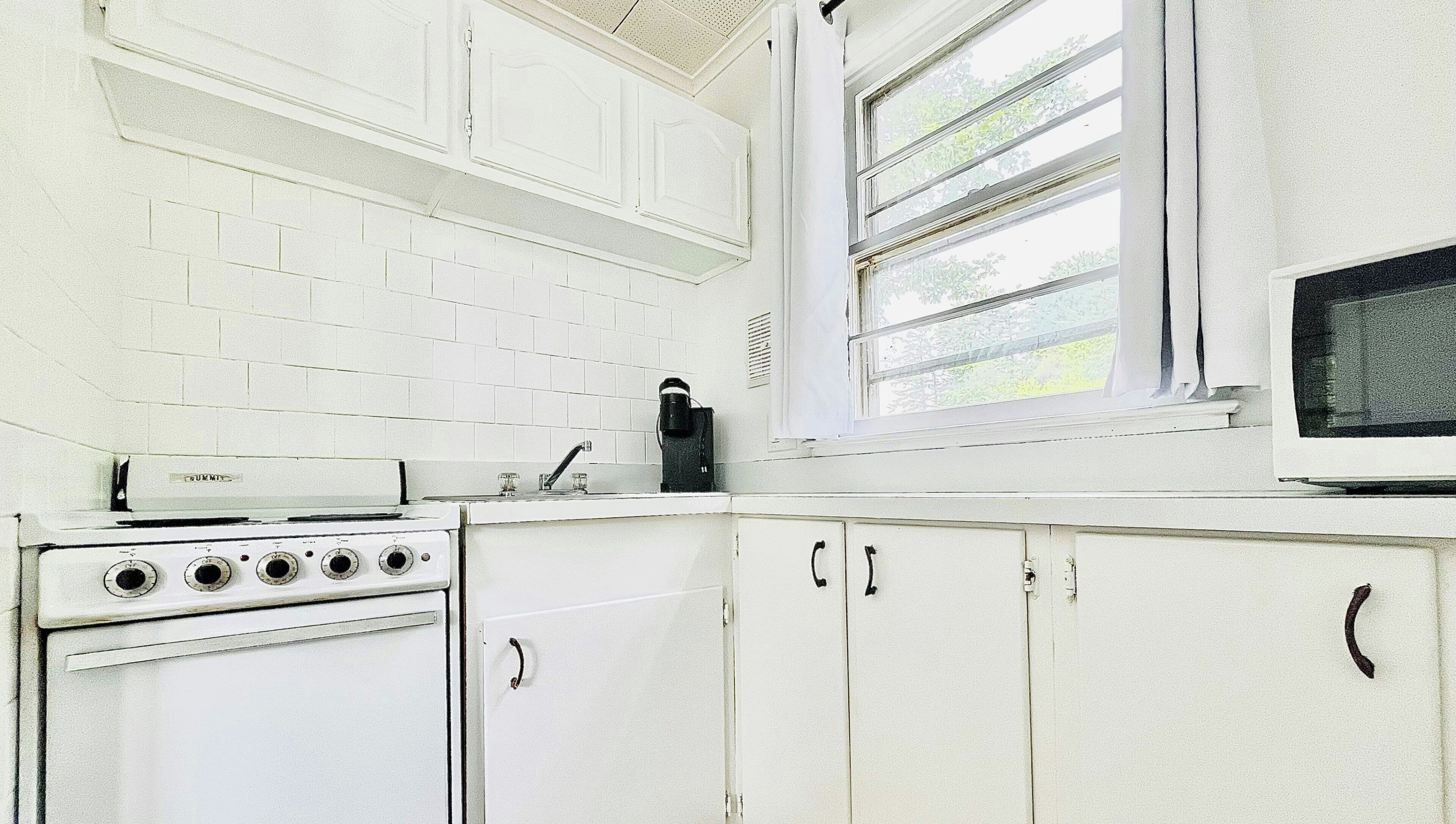 A kitchen with white cabinets, a stove, a coffee maker, and a window with blinds.