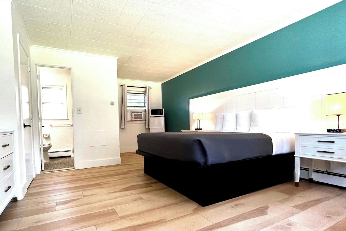 A bedroom with a bed, bedside table, lamp, and a view of the bathroom.