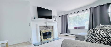 A modern living room with a fireplace, a flat-screen TV mounted above it, a large window with curtains, and a sofa with patterned cushions.
