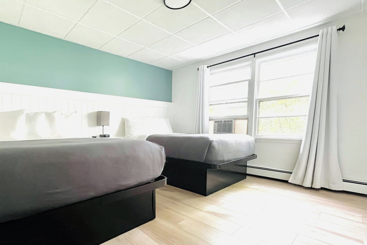 A hotel room with two beds, a window with curtains, a ceiling light, and a wall-mounted heater.