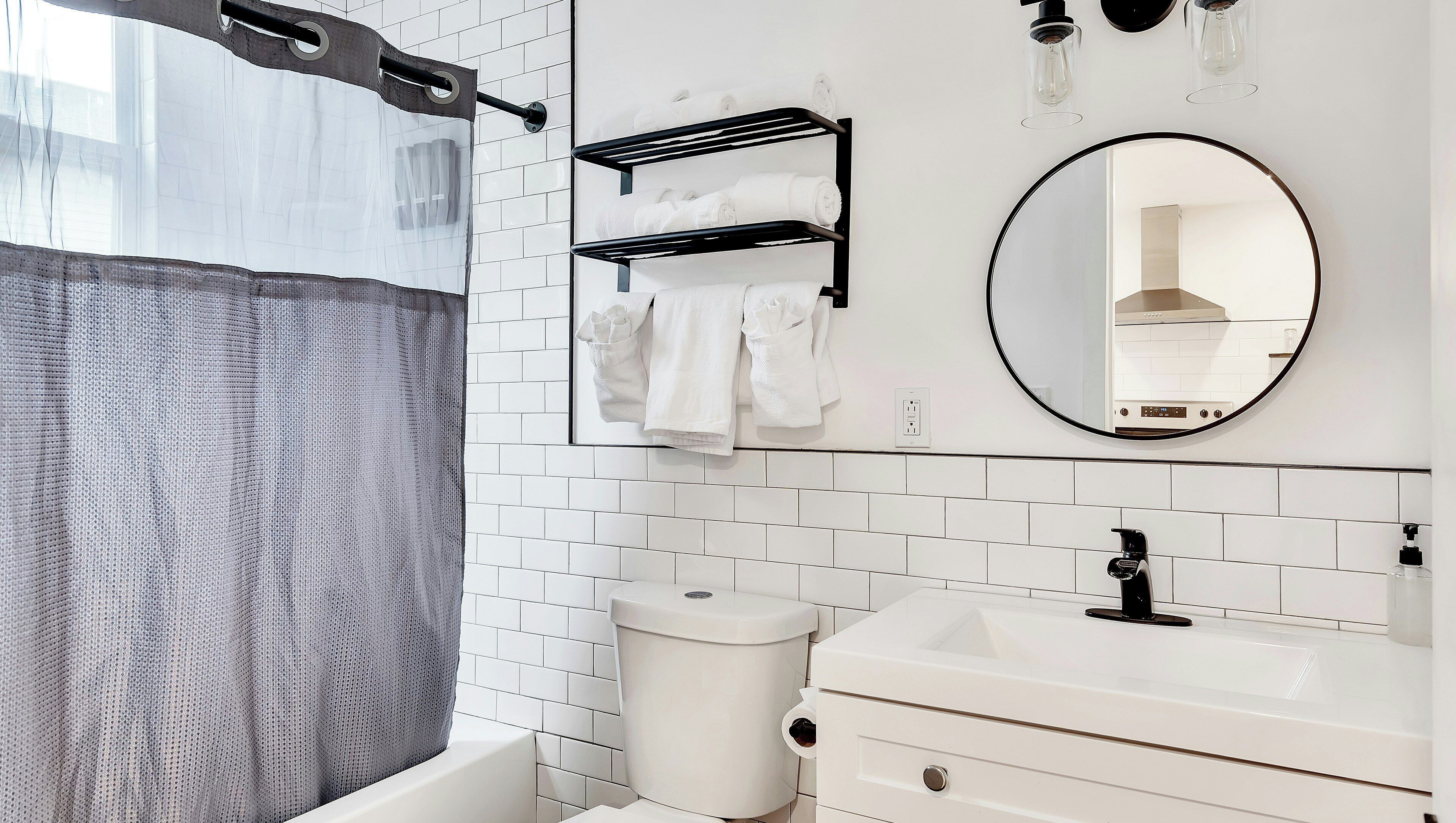 A modern bathroom with a bathtub, white tiled walls, a toilet, a sink, a mirror, and towel racks.