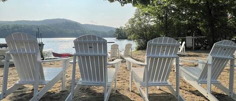 Ein Strand mit Adirondack-Stühlen, die auf einen See ausgerichtet sind, umgeben von Bäumen und einem Boot.