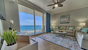 Un amplio salón con un gran ventanal que ofrece vistas a la playa, un ventilador de techo, un conjunto de sofás y una mesa de centro.