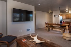 A living room with a flat-screen TV mounted on the wall, a wooden dining table with benches, and a kitchen area with wooden cabinets and a refrigerator. - Mountain Laurel Condo: Porch Views, AC, Close to Music on Main (Victor)