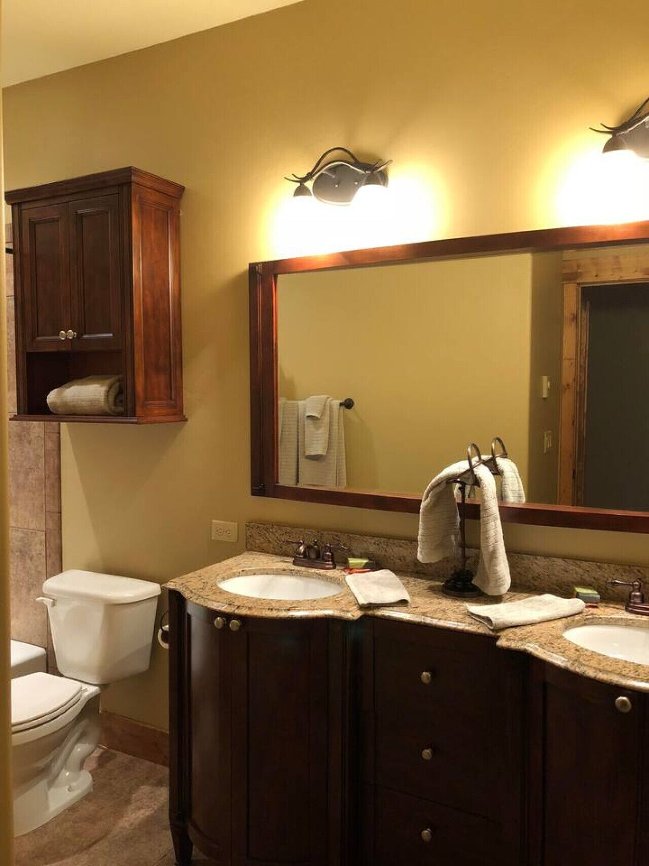 A bathroom with a toilet, two sinks, a vanity with a mirror, and a cabinet above the toilet.