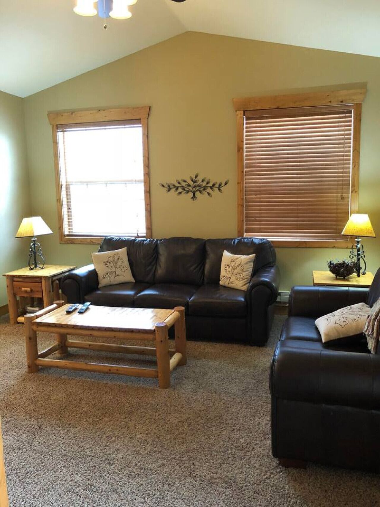 A living room with a high ceiling, two windows with blinds, a wooden coffee table, and a black leather sofa.