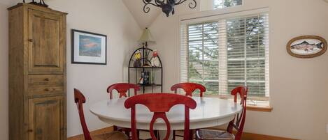 A dining area with a round table, red chairs, a wooden cabinet, a chandelier, and a window with blinds.