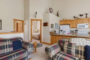 A cozy living room with plaid sofas, a wooden-framed doorway leading to a bedroom, and a kitchen area with wooden cabinets and a microwave. - Cozy 2 level townhome with scenic views - SR # 47 (Driggs)