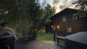 A wooden deck with a barbecue grill, a hot tub, and a cabin with windows lit from inside. - Windigo Lodge with Hot Tub (Victor)