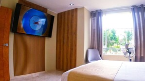 A hotel room with a bed, a TV mounted on the wall, a wooden panel wall, a window with curtains, and a fan. - SOL DE LUREN PUENTE BLANCO (Ica)