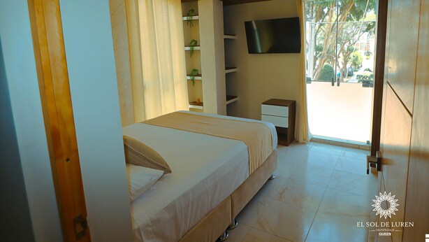 A bedroom with a bed, a TV mounted on the wall, and a window with a view of trees. - SOL DE LUREN PUENTE BLANCO (Ica)