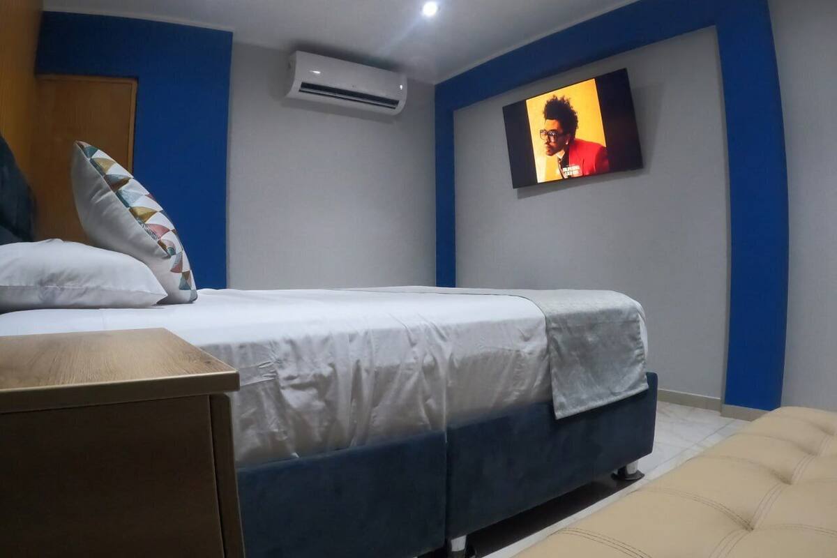 Une chambre d’hôtel avec un lit, un téléviseur fixé au mur, une table de chevet en bois et un fauteuil de couleur beige.