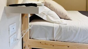 Une petite chambre avec un lit simple, un cadre en bois, une lampe blanche posée sur une étagère et une prise électrique au mur.