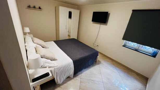 Une chambre à coucher comprenant un lit, une table de chevet, une lampe, une armoire, un téléviseur et une fenêtre avec des stores.