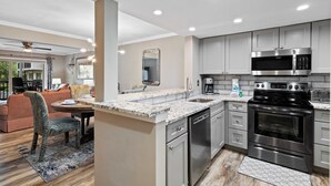 A modern kitchen with stainless steel appliances, a granite countertop, and a dining area with a sofa and table. - Beautiful Spacious Condo w/Pool walk to Beach, Celebration Park, Coligny Plaza! (Hilton Head Island)