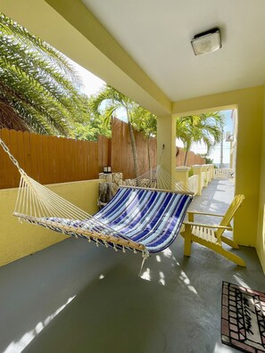 A hammock chair with a blue and white striped fabric, a wooden table with chairs, and a view of a wooden fence and palm trees.