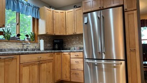 A kitchen with wooden cabinets, a stainless steel refrigerator, a coffee maker, and a window with a view of trees. - Beautiful Fall Foliage & Excellent Fishing on the Crown Jewel in the Northwoods (Mercer)
