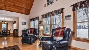 A spacious living room with two leather armchairs, a coffee table, and a rug. Large windows with blinds and curtains are visible. - Beautiful Fall Foliage & Excellent Fishing on the Crown Jewel in the Northwoods (Mercer)