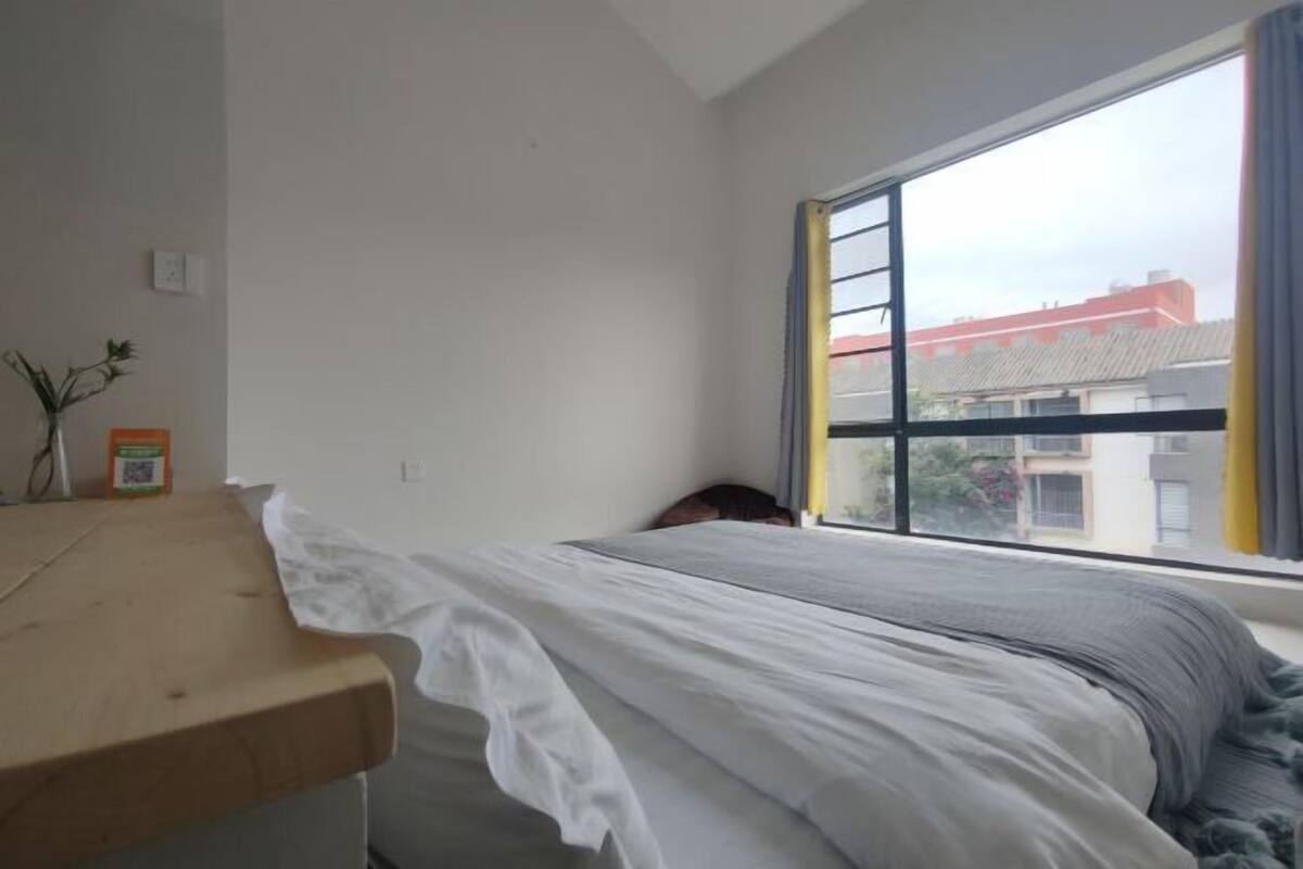 A bedroom with a bed, a wooden desk, and a window with a view of buildings.