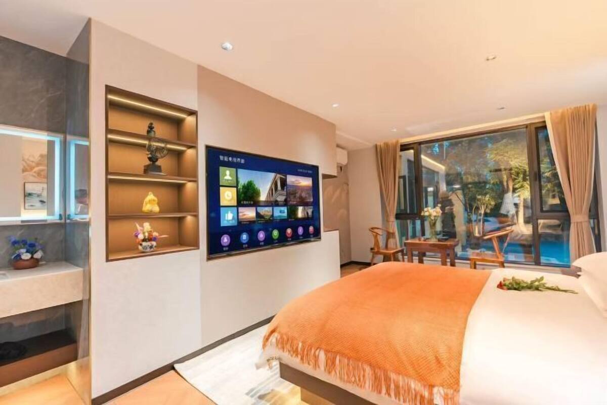 A modern hotel room with a large bed, a mounted TV, a desk with a chair, and a view of the outdoors.