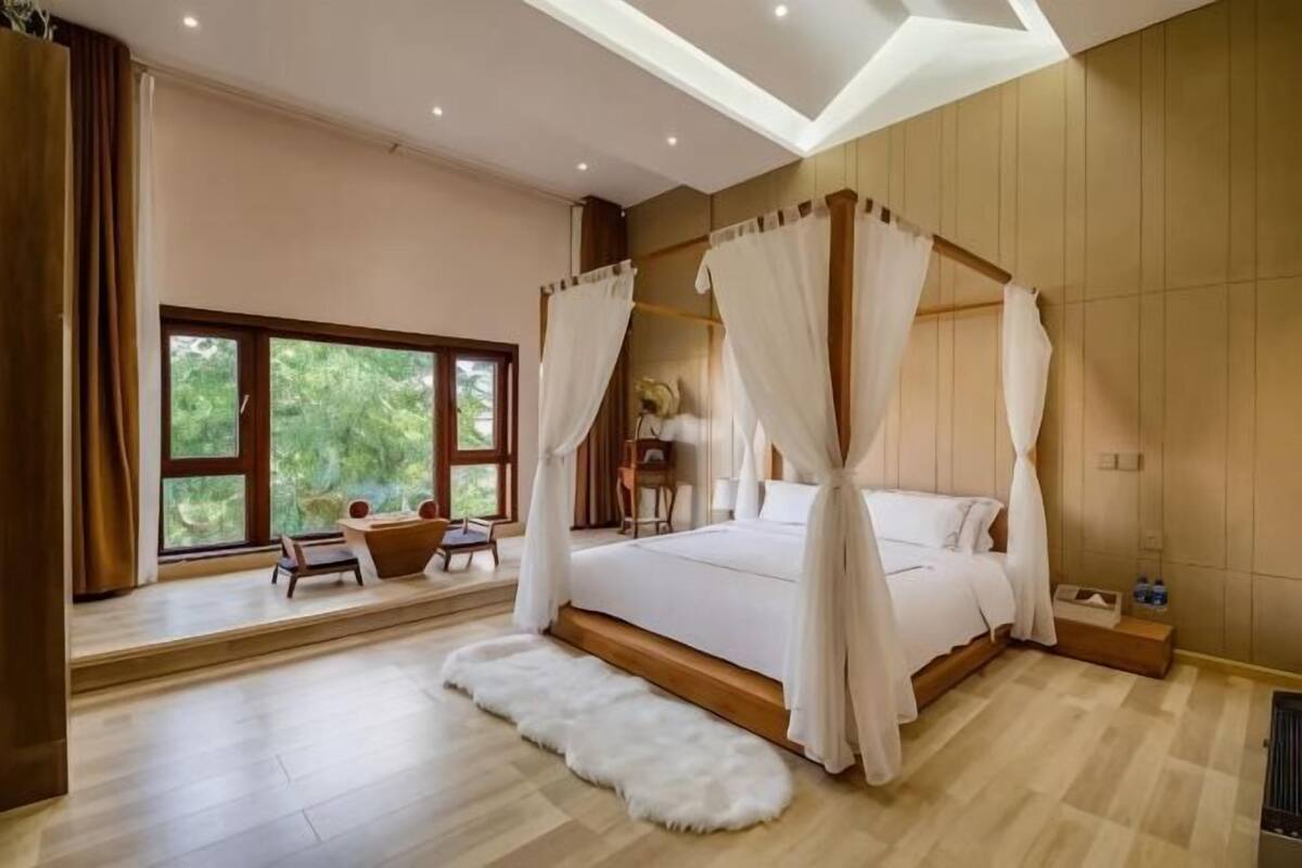 A bedroom with a four-poster bed, a large window, a small table, and a white fur rug.