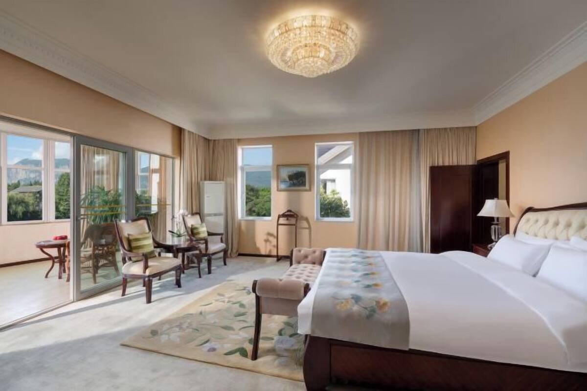 A hotel room with a large bed, a chandelier, a view of greenery through the windows, and a seating area with chairs.