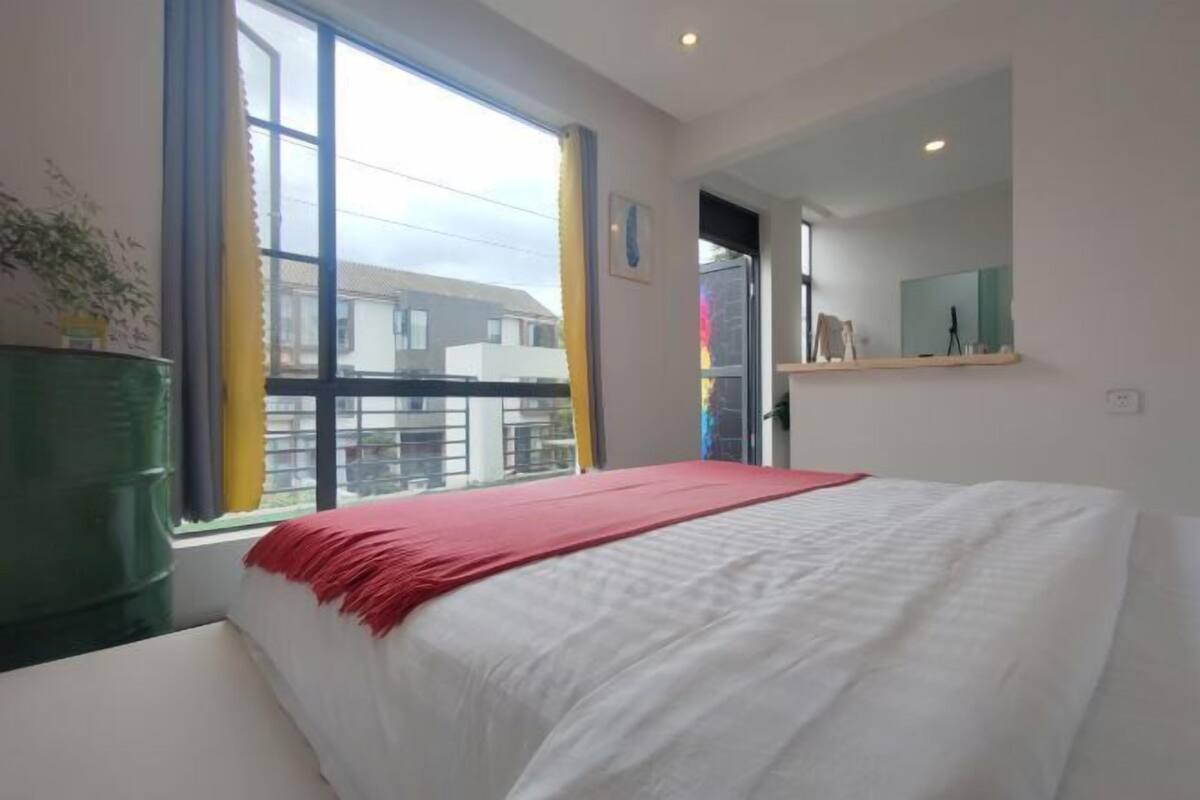 A bedroom with a large window, a bed with white and red bedding, a green chair, and a small plant.
