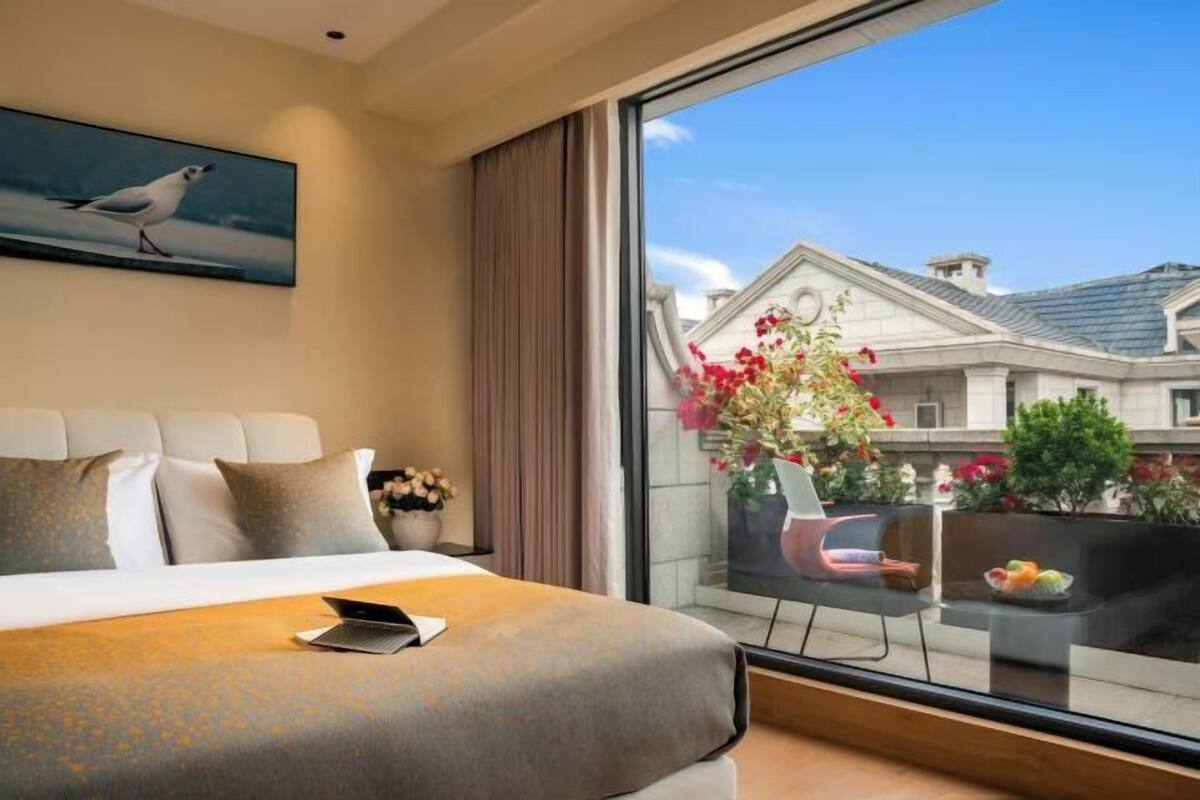 A hotel room with a large bed, a view of the city, a balcony with a table and chairs, and a painting on the wall.