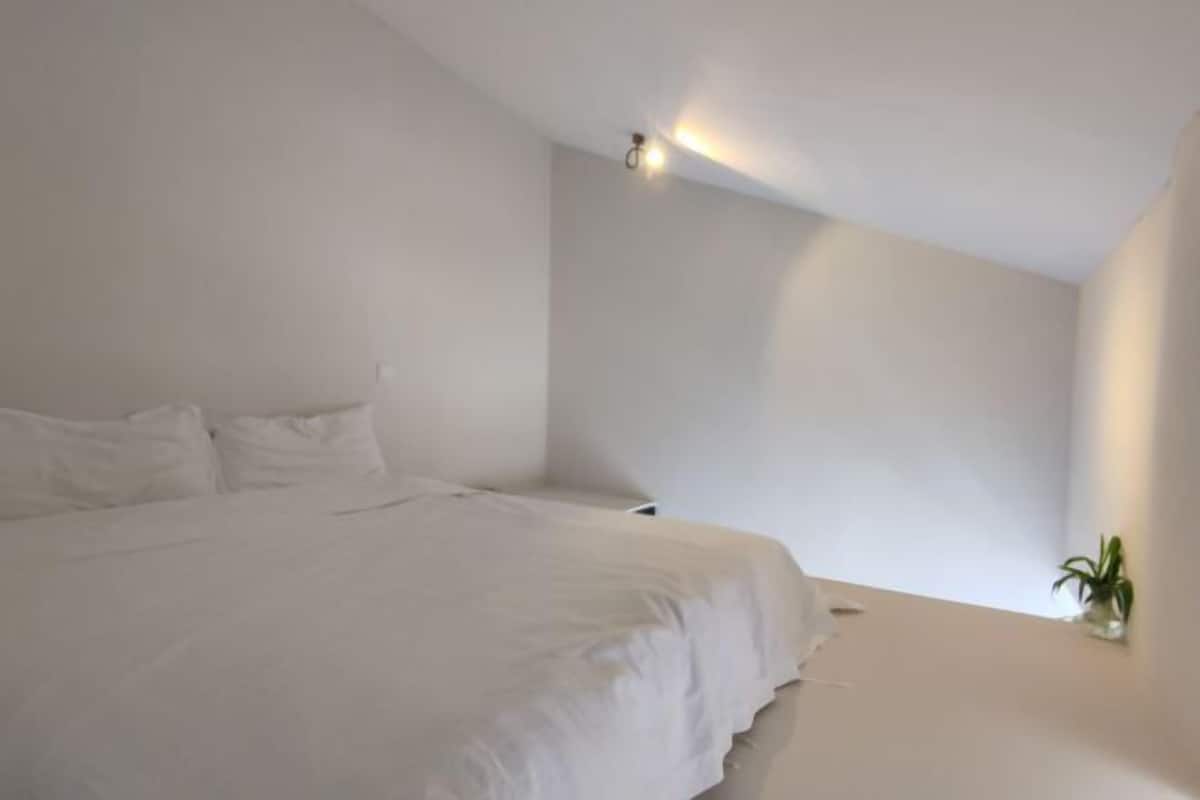 Quarto minimalista com uma cama, uma mesinha de cabeceira e um aplique na parede.