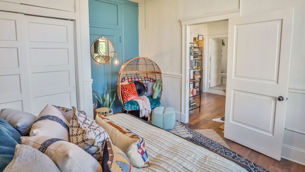A cozy room with a blue door, a wicker chair with cushions, a large mirror, and a bookshelf.