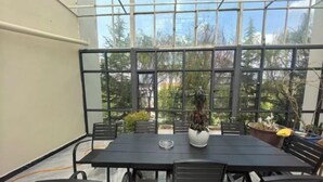 Un espace repas entièrement vitré, avec une table et des chaises noires, des plantes en pot et une vue sur les arbres et le ciel.