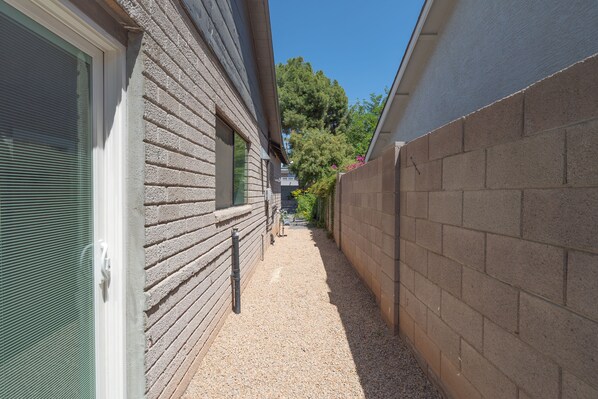 A narrow alleyway with a gravel path, a brick wall on one side, and a wooden building with a window on the other.