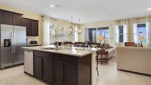 A modern kitchen with dark cabinetry, a stainless steel refrigerator, and a central island with a sink. - Sunset Shores Villa at Solterra Resort | Luxurious 7BR Lake View Home with Priva (Davenport)