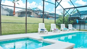 Une piscine entourée de quatre chaises longues blanches, une terrasse fermée par des baies vitrées et, en arrière-plan, une vue sur des maisons et une colline verdoyante.