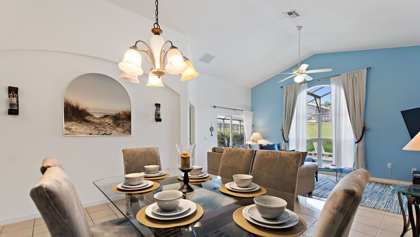 Un coin repas avec une table en verre dressée pour six personnes, entourée de chaises rembourrées. Un ventilateur de plafond et un luminaire suspendu sont visibles au-dessus de la table.