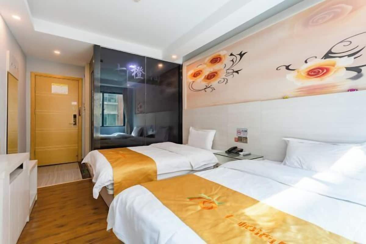 A hotel room with two beds, a wooden floor, a large floral artwork on the wall, and a door to the left.