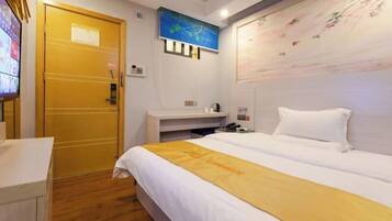 A hotel room with a bed, a wooden door, a wall-mounted TV, a painting, and a small table with a phone and a cup.