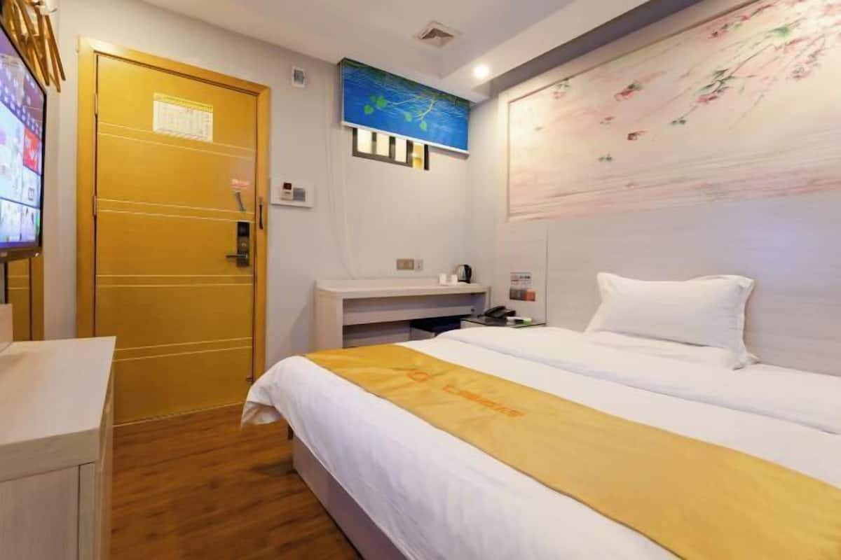 A hotel room with a bed, a desk, a wooden door, a wall-mounted TV, and a painting.