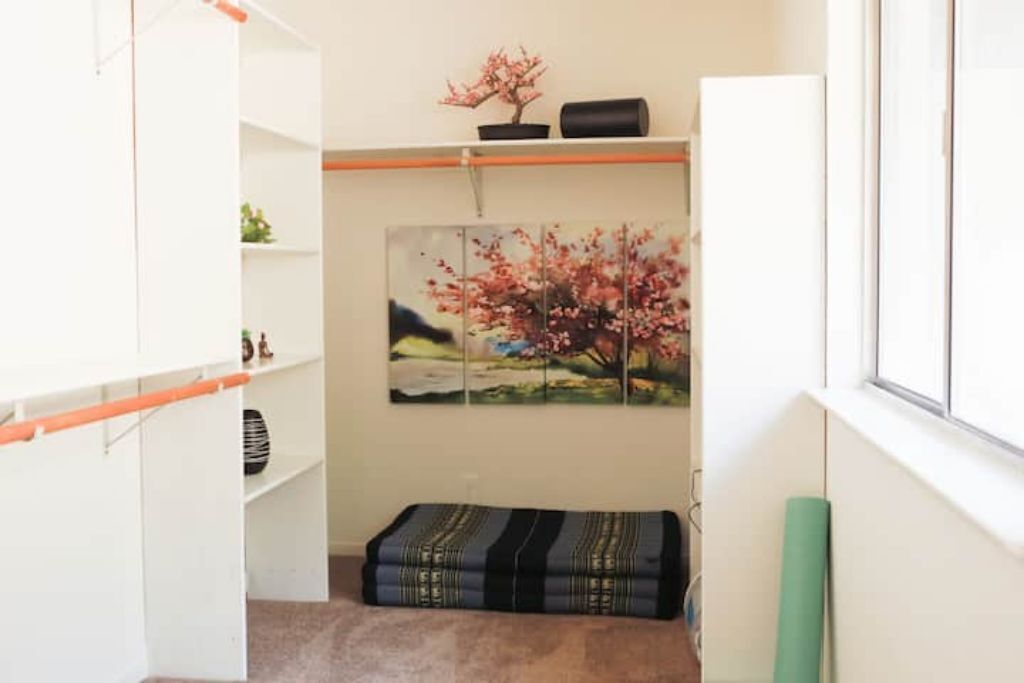 Una habitación con pared blanca, un cuadro de un árbol de cerezo, un banco con estampado negro y dorado, y un armario blanco.