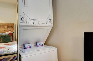 Eine Waschmaschine mit Frontlader, Waschmittelkartons auf der Oberseite, ein Bett mit einer Steppdecke und ein Fernseher an der Wand.