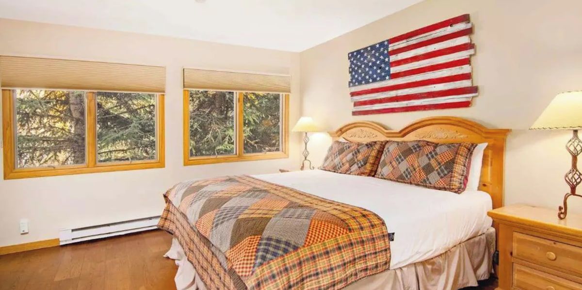 Een slaapkamer met een bed, twee ramen, een aan de muur hangende Amerikaanse vlag en een nachtkastje met een lamp.