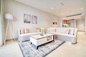 Un salon moderne avec un canapé blanc, une table basse en verre et un tapis à motifs bleus. Deux tableaux encadrés ornent le mur, et une lampe sur pied est placée dans un coin.
