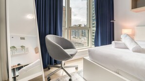 A modern hotel room with a bed, a desk, a chair, a window with curtains, and a city view. - Alluring 2BR at Al Majara 1 Dubai Marina by Deluxe Holiday Homes (Dubai)