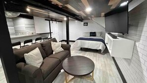 Een moderne hotelkamer met een bank, een ronde salontafel, een bed met een blauwe spreideken, een witte nachtkastje en een flatscreen televisie aan de muur.