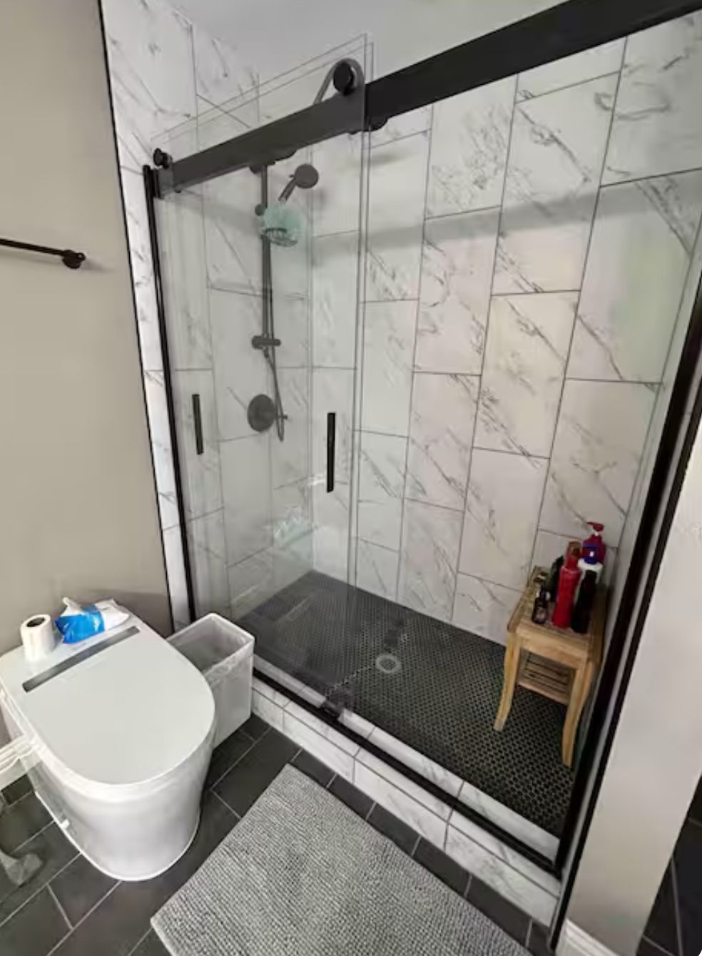 Moderni kylpyhuone, jossa on lasiseinällä varustettu suihku, wc ja pieni puinen jakkara, jonka päällä on hygieniatarvikkeita.