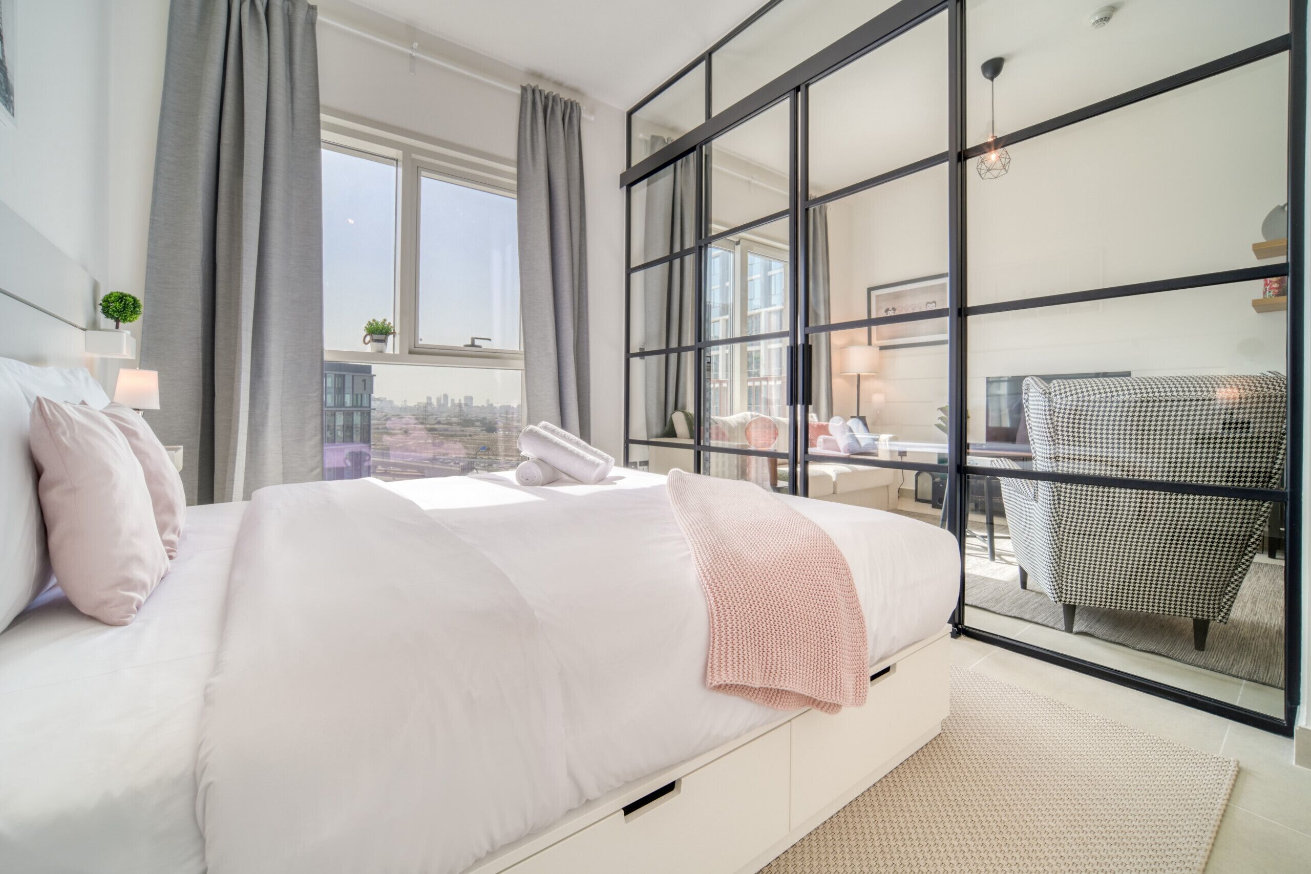 Une chambre moderne avec un grand lit, une vue sur la ville grâce à de grandes fenêtres du sol au plafond et un balcon avec un fauteuil.